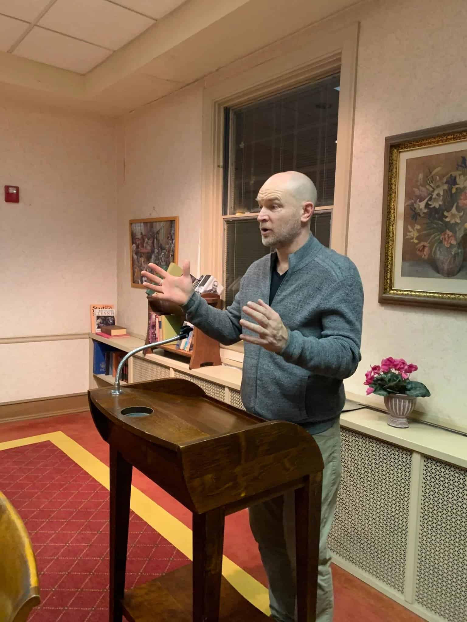 Speaker delivering a talk from a podium in a warmly lit room, addressing personal development or mental health topics.