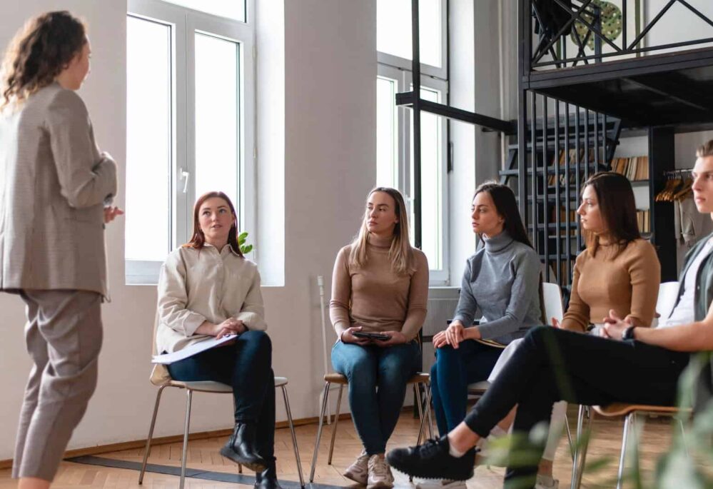 Facilitator leading a small group therapy or coaching session in a modern, well-lit space focused on mental wellness and emotional growth.
