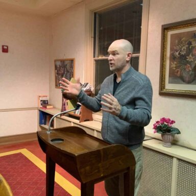 Speaker delivering a talk from a podium in a warmly lit room, addressing personal development or mental health topics.